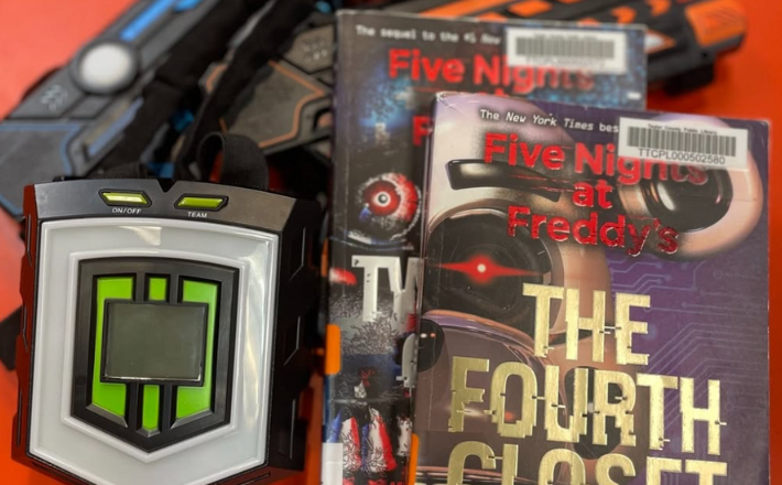 Library Laser Tag promotion with laser tag gear beside the novel The Fourth Closet